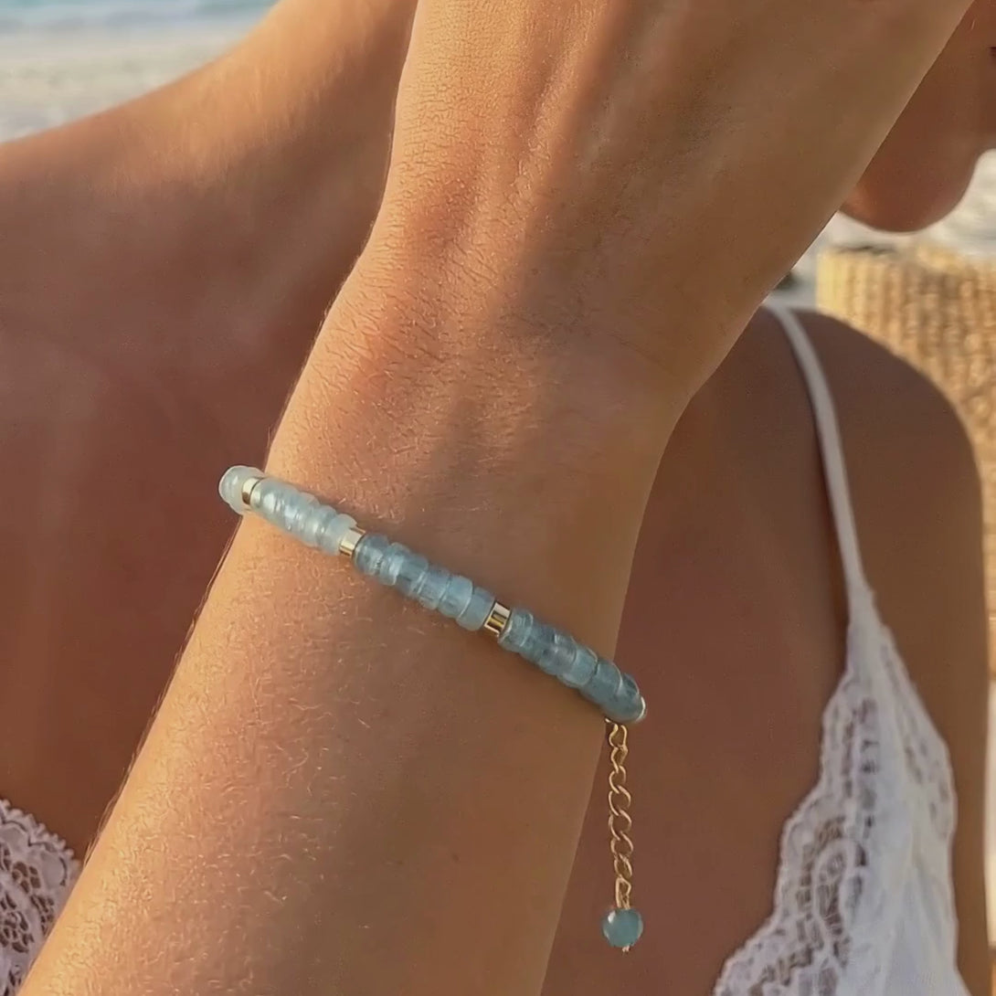 Video showcasing a woman in a flowy white dress wearing a natural aquamarine 14K gold bracelet walking by the beach.